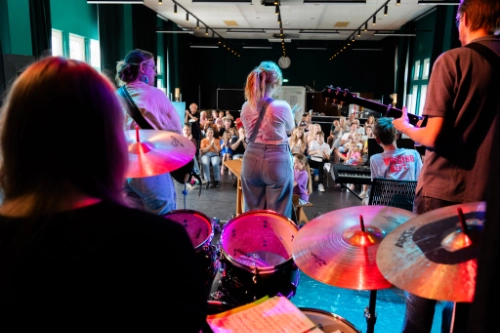 Bandje gefotografeerd van achter de drummer. Links staat een vrouwelijke gitarist, rechts een mannelijke. En de zangeres staat in het midden.